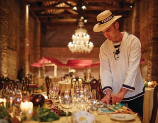 A man dressed in a traditional navy and white gondolier smock and straw hat sets a table ready for a party in a grand hall