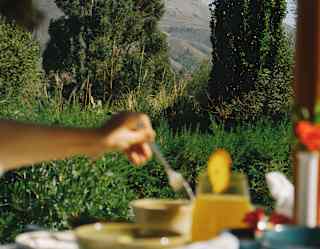 In soft-focus foreground, a woman stirs her coffee at an outdoor breakfast table, with a backdrop of garden trees and hills.