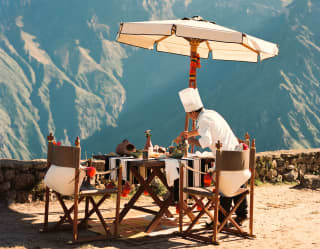 A chef arranges breakfast for two on a table with a striped cloth and a parasol at Tapay, with breathtaking mountain views.