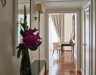 In the foreground a vase of bougainvillea blushes dark pink. Polished floors lead along a corridor to a light filled room