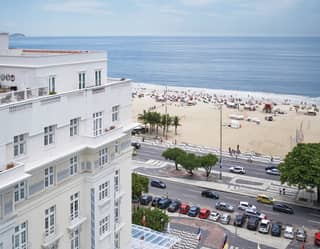 Calm blue sea laps the bustling Copacabana Beach. People stroll the sidewalk. Its pattern is repeated on the hotel's forecourt