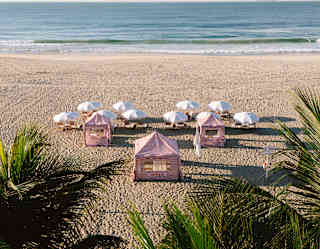 Three red-striped cabanas and ten pairs of loungers shaded by white parasols face the sea in a view of the beach over palms.
