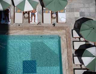 Aerial shot of a corner of the sun-rippled pool bordered by loungers, where a tanned man relaxes, face obscured by a parasol.