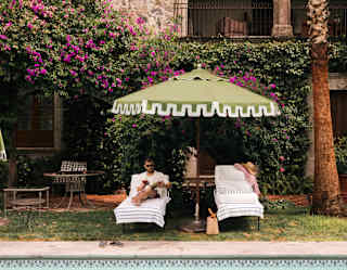Two pool-side sun loungers recline beneath a green parasol, with a hat and shirt on one, and a man reading on the other.