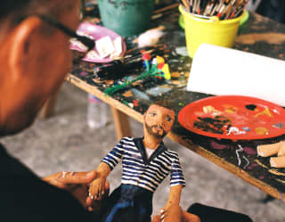 Artist Hermes Arroyo holds a small mojiganga puppet with a stripy blue top at a workshop table busy with paint and brushes.