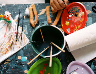 Two pairs of mojiganga arms wait to be painted on a workshop table with kitchen towel, water and brushes, seen from above.