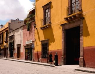 Two women dressed in black walk by a large terracotta and yellow building with a huge doorway in San Miguel de Allende.