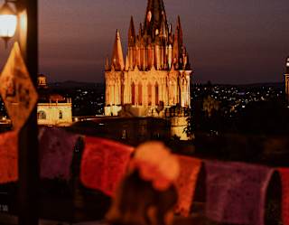Parroquia de San Miguel Arcángel at night, illuminated, with a glowing cross on the top, viewed from the rooftop bar.