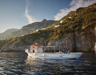 Small white motor boat sailing alongside the rugged cliffs of the Amalfi Coast