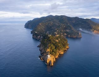 Aerial view of Portofino lighthouse at the tip of the rocky forest-covered peninsula, surrounded by deep blue ocean.