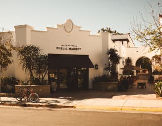 Kodachrome-style image of the white Santa Barbara Public Market building, with a small outdoor café and potted palms.