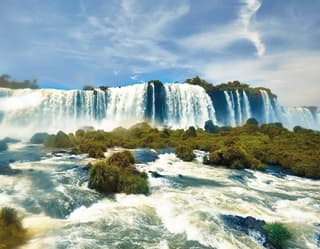 Low angle view from the grassy rock-filled water, as the Iguaçu River tumbles over a wide ledge in impressive cataract falls.