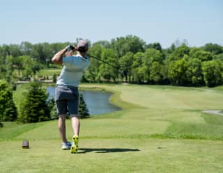 in a verdant golf course, a man in white shirt and dark shorts watches his airborne ball, following a swing, seen from behind.