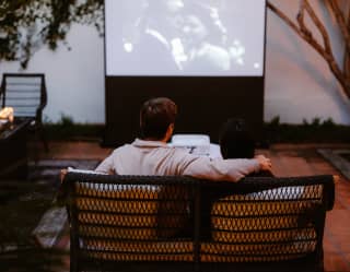 A couple cuddle on a sofa watching a black and white film at Al Fresco Movie Night on Oak Tree Suite Patio, seen from behind.