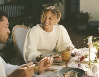 A woman in a white jumper with curly hair smiles as a male friend talks during a meal at a pretty table with vibrant plates.