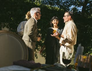 Two men and two women chat in the gardens of the Oak Tree Suite, standing in sunshine, viewed over a shaded dining table.