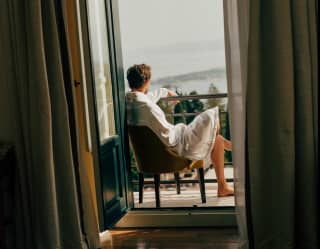 A man in a white robe relaxes in a chair on a balcony, gazing at the views, seen from inside the suite through open doors.
