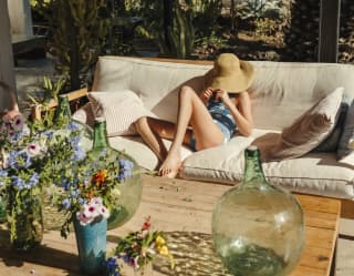 A girl sinks into the cream cushions of a large outdoor sofa, her whole head swallowed by a straw hat. Flowers fill the table
