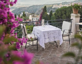 Bougainvillea tumbles down, framing the view of a lace topped terrace dining table for two, with Mount Etna rising behind
