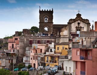 A street view in Forza D'Agrò shows the bell tower and roofline of Chiesa della Santissima Annunziata above a pastel terrace