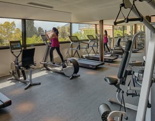 A woman in black leggings and pink t-shirt looks out at the view of Naxos Bay while working out on a cross-trainer in a gym