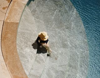 Birds-eye-view of a guest in a wide-brimmed sun hat lounging in the shallow water of an outdoor pool