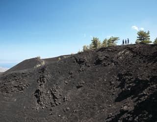 Tourists standing on the dark, rocky slopes of Mount Etna under blue skies