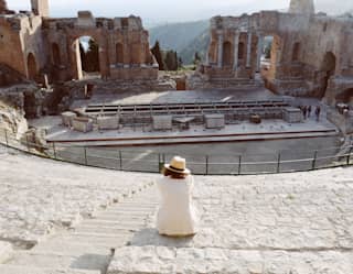 A women in a white jacket and panama hat sits on the stone steps of the Greek theatre as technicians build a stage below