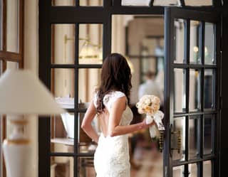 Bride in a fitted wedding dress walking through floor to ceiling glass-panel doors