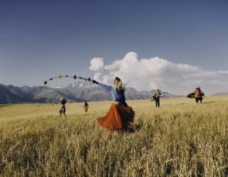 A woman in a red skirt, blue top and traditional headdress, waves floral decorations in an open grass field with four others.