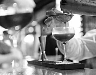 Black and white close-up of a bartender straining a drink from a silver cocktail shaker through a sieve into a glass.