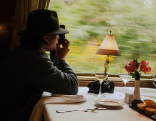 A guest in a fedora hat photographs the passing landscape through the window, seated at a dining table in the restaurant car.