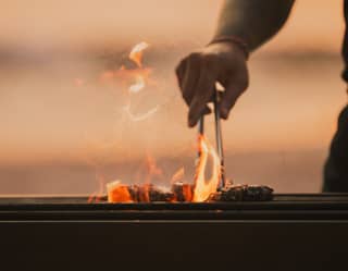 A chef turns morsels with tongs as flames and tendrils of smoke lick the air, seen in a close-up detail of a sunset barbeque.