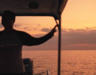 Image of a man as he holds the canopy support of the Dedari boat, viewed in silhouette against the ocean's sunset ripples.