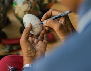 A local man decorates a painted white egg with a fin-tip pen, in an activity detail viewed over the artist's shoulder.