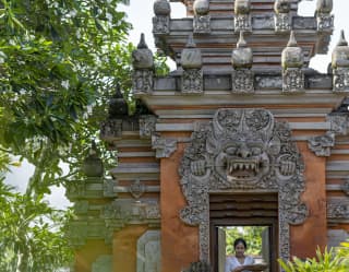 A woman walks through a traditional Kori Agung Balinese gate, which is orange with a pagoda-style pyramid roof with carvings.
