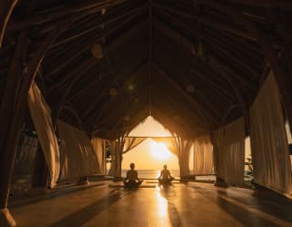 Two guests in lotus pose face the sea and sun, which bathes the yoga pavilion in honey light and shadows, seen from behind.