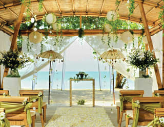 Bamboo pergola over a beach set with rows of chairs and an aisle for a wedding