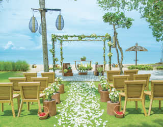 On a lawn overlooking a beach, twelve chairs flank a path of flowers to a floral-festooned wedding altar with a sea backdrop.