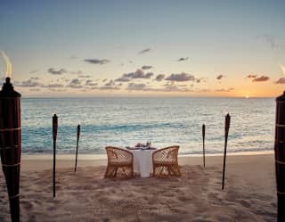 Private beach table at sunset with fire-lit torches framing a sea view