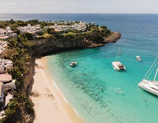Four boats float in Baie Longue's sapphire waters, sheltered by a headland dotted with resort residences, in an aerial view.