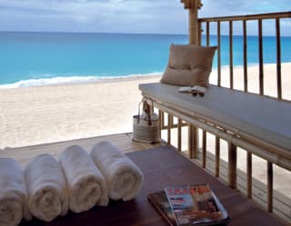 A straw-roof private cabana is furnished with cushioned benches, hardwood table and rolls of fluffy white towels