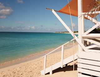 A white wooden beach hut with an orange canopy stands on sandy shore next to turquoise sea under a clear blue sky.