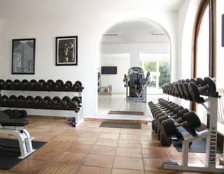 Rows of free weights sit neatly on tiered stands. An archway opens onto the white tiled fitness room, revealing the leg press