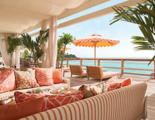 A large sofa with coral cushions fills the foreground. Behind, tables await diners. On the balcony loungers overlook the sea