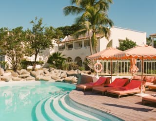 Pink lounge chairs recline under parasols at the edge of the tropical pool with sweeping in-pool steps and rockery border.