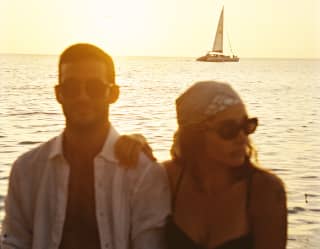 A man in dark glasses and a woman with a headscarf and shades sit in honey sun as a boat sails across the ocean behind.