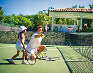 Tennis coach kneeling beside tennis net while little girl practices serve