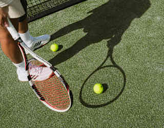 A tennis player stands at the net, with one of three grounded balls resting on the shadow of the racquet cast onto the court.