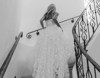 A black and white image of a bride in a flowing lace gown climbing white marble spiral stairs with ornate black ironwork railings